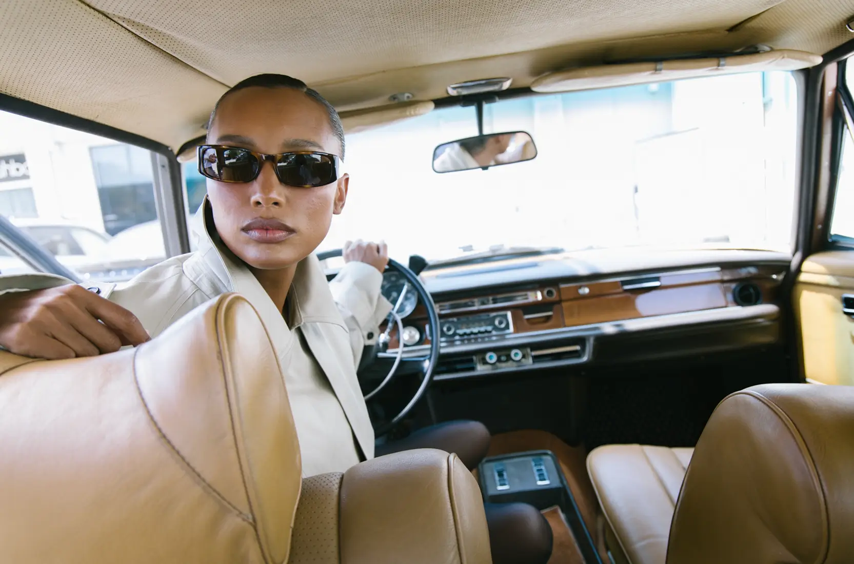 RHEA Creative Woman wearing sunglasses and a light jacket sits in the driver’s seat of a vintage car, looking over her shoulder toward the backseat. Luxury Branding